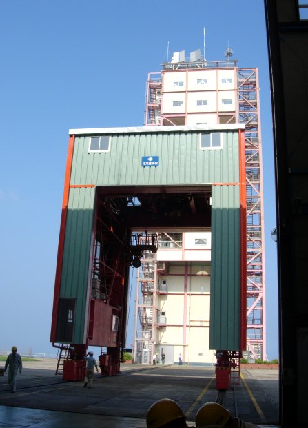 A large house-shaped crane on widely spaced railroad tracks is silhouetted against a bright blue sky, as the 10-story launch tower looms in the background (51K JPEG)