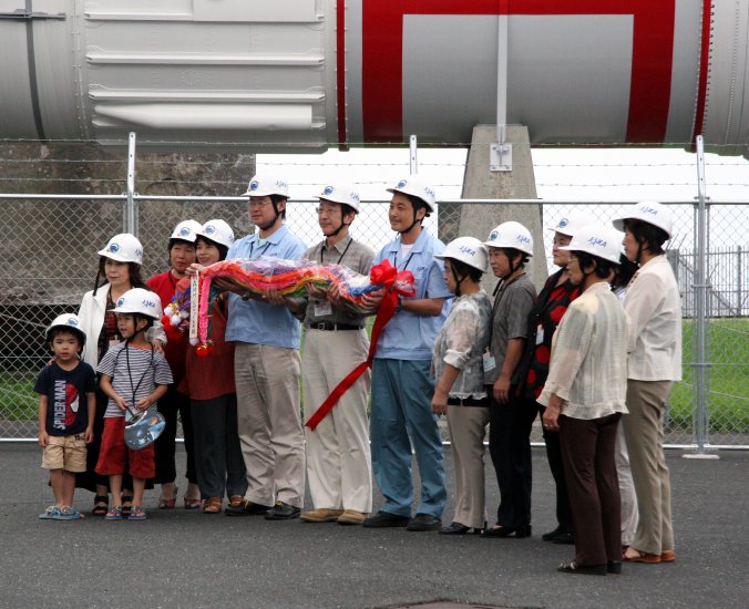 Inoue-sensei, looking most dapper in a white hardhat and flanked by coworkers, townspeople, and cute children, receives a long, colorful, beribbonned gift. (92K JPEG)