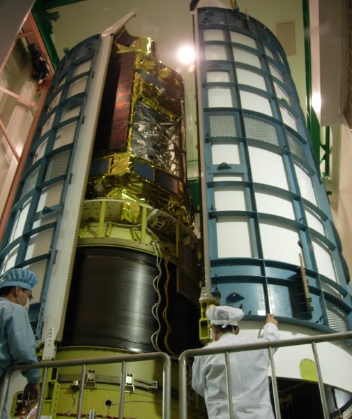 The spacecraft towers over us, mounted on a sleek black cylinder which is the third stage pressure vessel.  One half of the cylindrical nose fairing (with conical top) is in place, as the other half is being slid toward it. (73K JPEG)