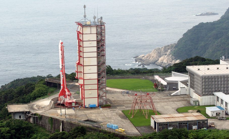 The rocket, attached to its fully saturated launcher, towers on the left side of the flat launch area, as the foliage-draped land drops away behind to the sea. (120K JPEG)