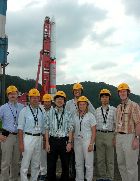 Eight happy people, lovingly attired in their bright yellow hardhats, pose before the towering might of the M-V-6 rocket. (87K JPEG)