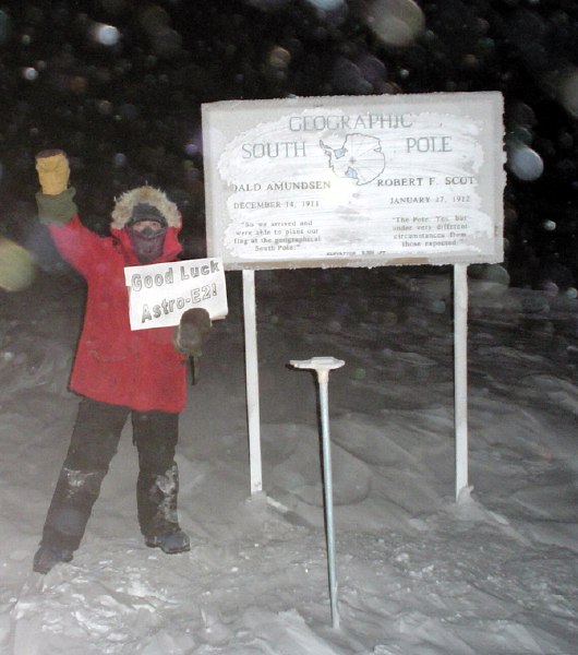 Flakes of snow and fog slightly blur the image of a parka-clad person with fist raised high, holding a 'Good Luck Astro-E2'; sign in the other hand, directly in front of the sign declaring 'Geographic South Pole' (64K JPEG)