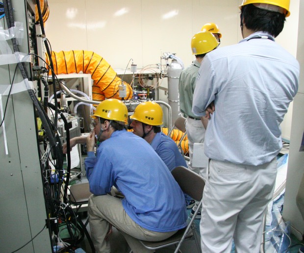 Two blue-smocked people in yellow hardhats sit looking carefully at a computer out of frame to the left, as three other people, also in yellow hard headwear stand and discuss other issues. (94K JPEG)