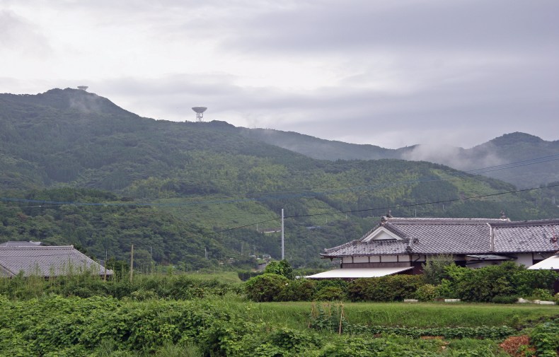 Lush greenery fades away from the graceful lines of traditional Japanese architecture to the horizon high above, flecked with two parabolic dishes, both in the up-facing stowed position. (100K JPEG)