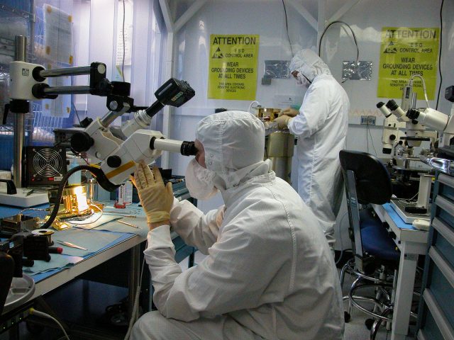 The shiny gold FEA is dwarfed by the binocular microscope through which the clean-suited John examines it.  (70K JPEG)