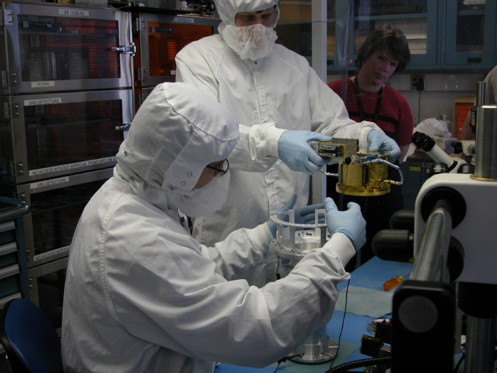 Two clean-suited people hold the shiny gold FEA and the metallic cylinder of the ADR. (60K JPEG)