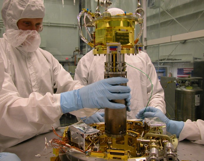 Gloved hands gently guide the 7 cm diameter ADR cylinder into a hole, not actually visible amid the mass of plumbing and mechanical structure that makes up the top of the insert.  (91K JPEG)