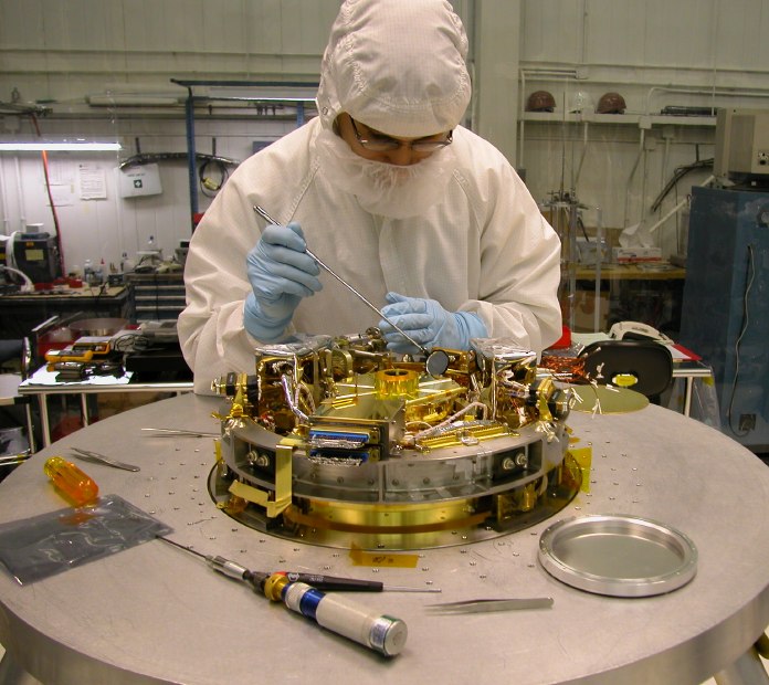 The tangly mass of shiny gold bits comprising the top of the helium insert protrudes from the much larger aluminum disk of the support stand, as a clean-suited engineer examines it closely with a small mirror on a long stick. (100K JPEG)